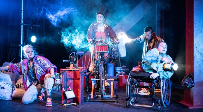 Four women on stage, one is riding a bike, one is kneeling down, two are shining torches