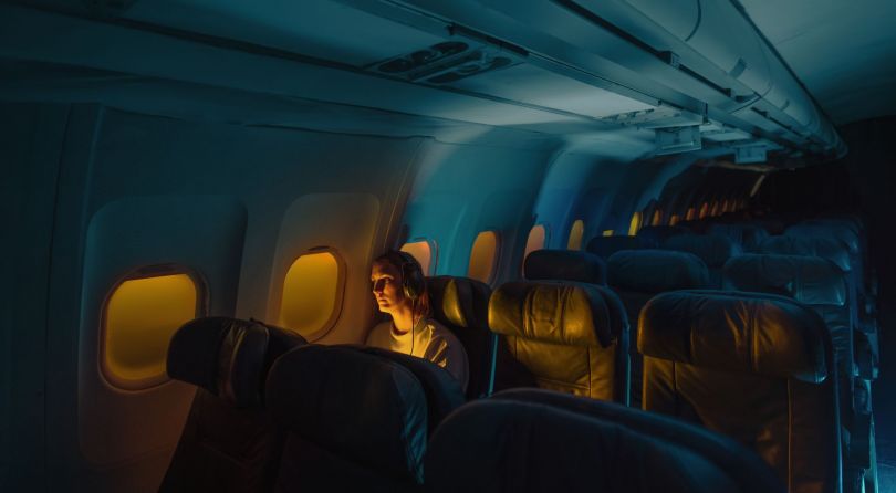 A woman sitting looking out of the window of an airplane. The seats around her are empty. She is wearing headphones.