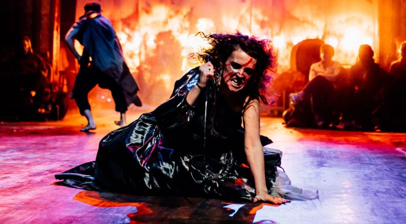 Woman kneeling on floor with a dagger in her hand, about to strike down. A figure is behind her and there are flames in the background